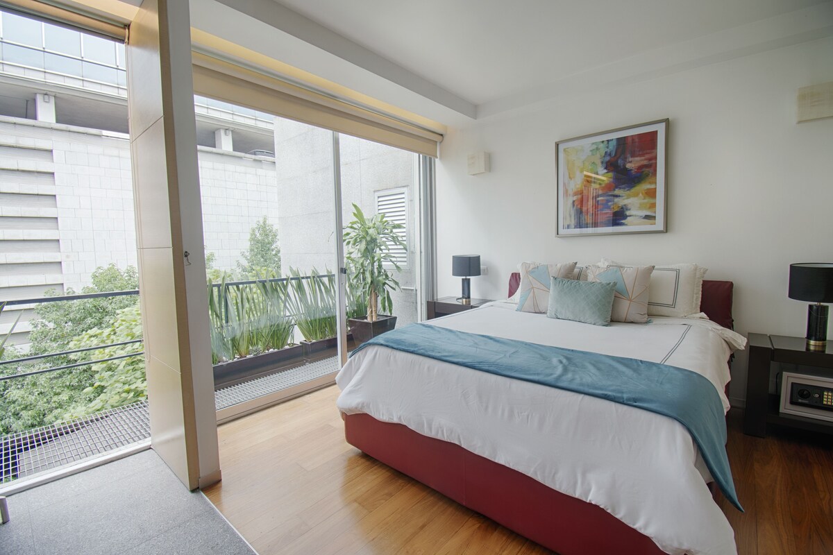 A bedroom with a large bed, a window with a view of greenery, a painting on the wall, and a nightstand with a lamp.