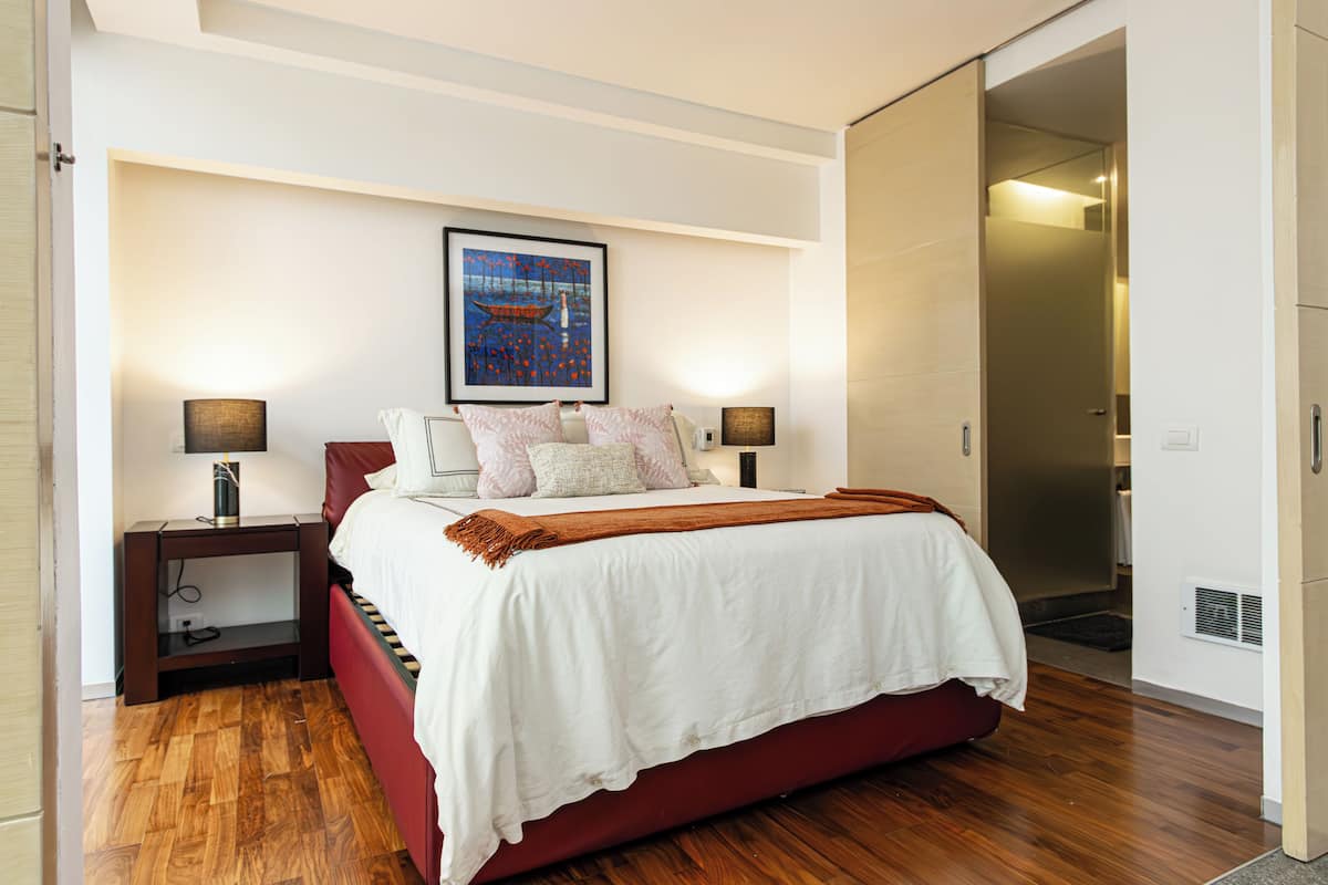 Une chambre moderne avec un grand lit, une table de chevet avec une lampe, une œuvre d’art encadrée au-dessus du lit et une porte coulissante donnant sur une autre pièce.