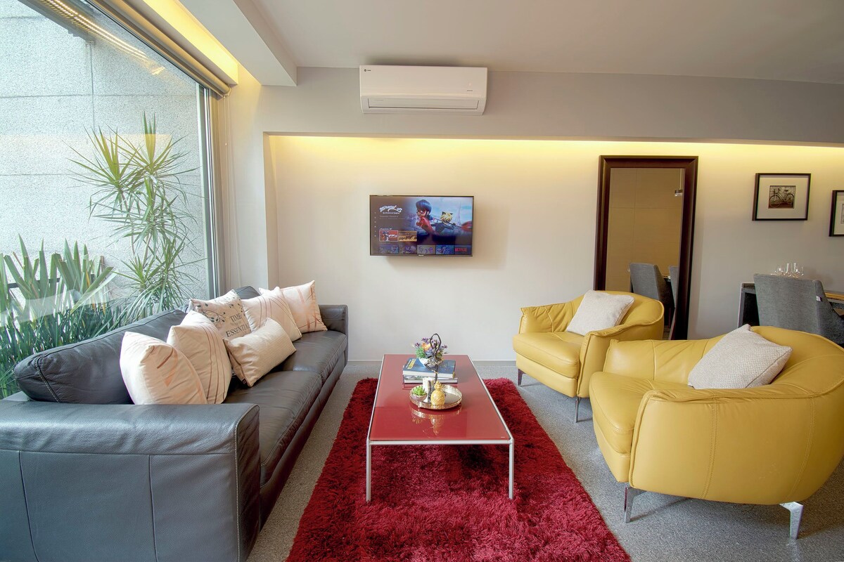 A modern living room with a grey sofa, yellow armchairs, a red coffee table, and a TV on the wall.