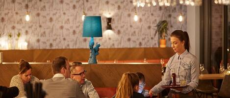 Interior de restaurante con barra de madera, luces colgantes y un fondo de papel tapiz estampado.