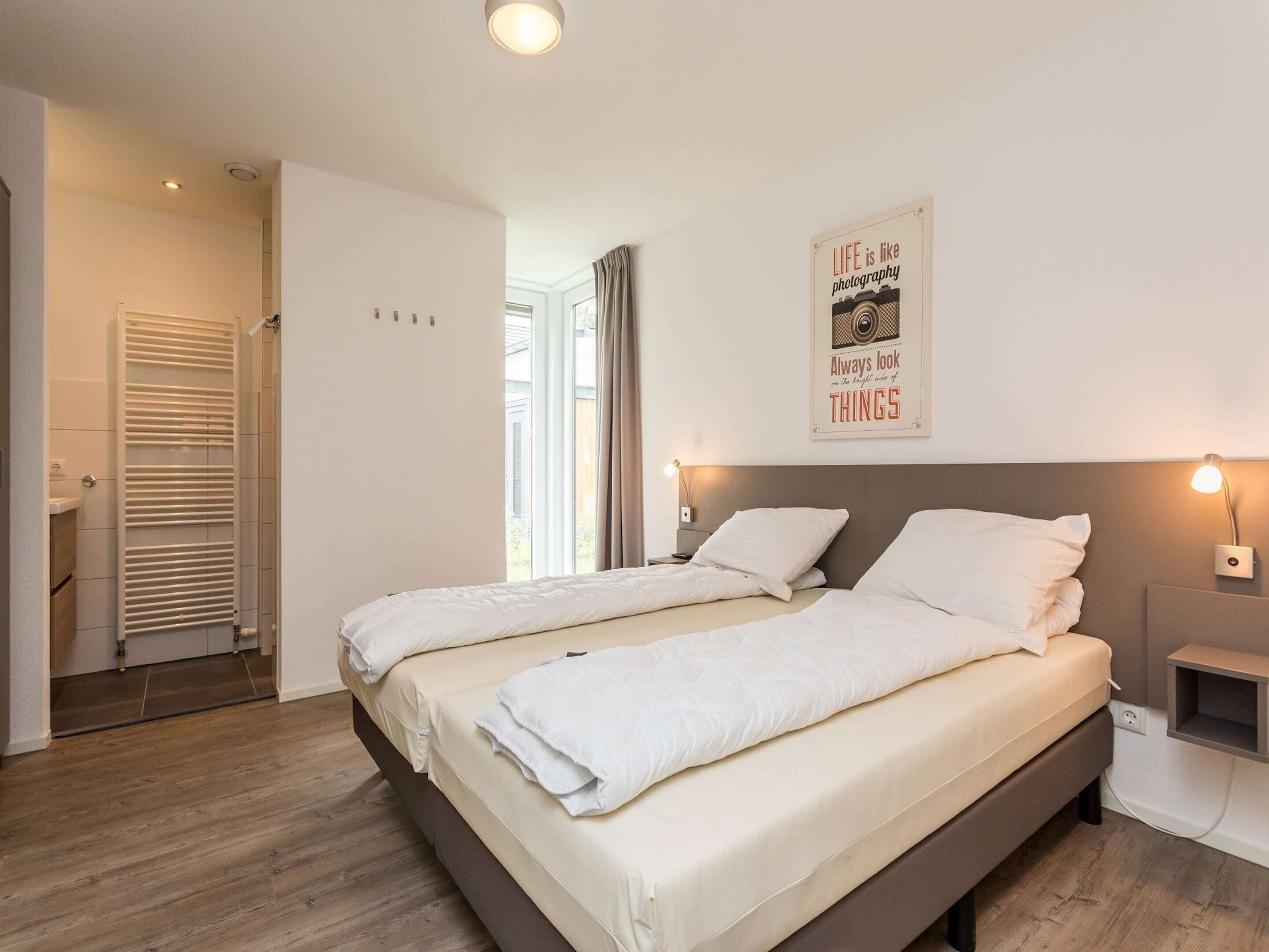 A modern bedroom with two beds, a large window, a radiator, and a motivational poster on the wall.