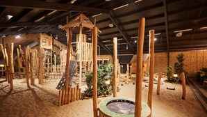 Une structure de jeu en bois comprenant un mur d’escalade et une balançoire, installée dans un espace intérieur sablonneux.