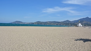 Μια παραλία με ένα λευκό κτίριο, βουνά στο βάθος και έναν καθαρό, γαλάζιο ουρανό.