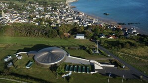An aerial view of a coastal town with a large, modern building with a dome structure, surrounded by residential houses and a beach. - Denneville Coastal Escape With Garden (Port-Bail-sur-Mer)