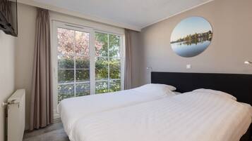 A hotel room with two beds, a large window with curtains, a round mirror on the wall, and a radiator.