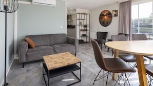A modern living room with a grey sofa, a wooden coffee table, and a dining area with a round table and chairs. There is a kitchen visible in the background, and a large window with a view of a body of water. - Luxe Chalet w/ Sauna (Hoge Hexel)