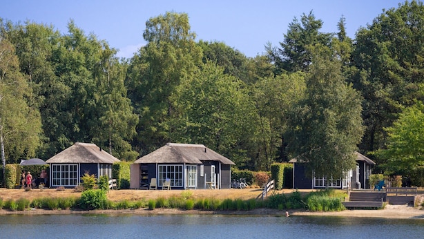 A lakeside property with thatched-roof cottages, a swimming area, and a forested backdrop. - Luxe Chalet w/ Sauna (Hoge Hexel)