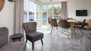 A modern dining area with a table and chairs, large windows with a view of a lake, and a flat-screen TV mounted on the wall. - Luxe Chalet w/ Sauna (Hoge Hexel)