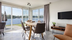 A modern dining area with a large table and chairs, a television mounted on the wall, and a view of a lake through large windows. - Luxe Chalet w/ Sauna (Hoge Hexel)