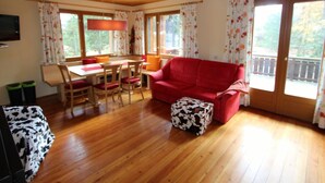 Un salón acogedor con un sofá rojo, una mesa de comedor de madera y sillas. Hay ventanas grandes con cortinas florales, un ventilador de techo y un televisor en la pared.