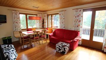 Ein gemütliches Wohnzimmer mit einem roten Sofa, einem Holzesstisch und Stühlen. Es gibt große Fenster mit floralen Vorhängen, einen Deckenventilator und einen Fernseher an der Wand.