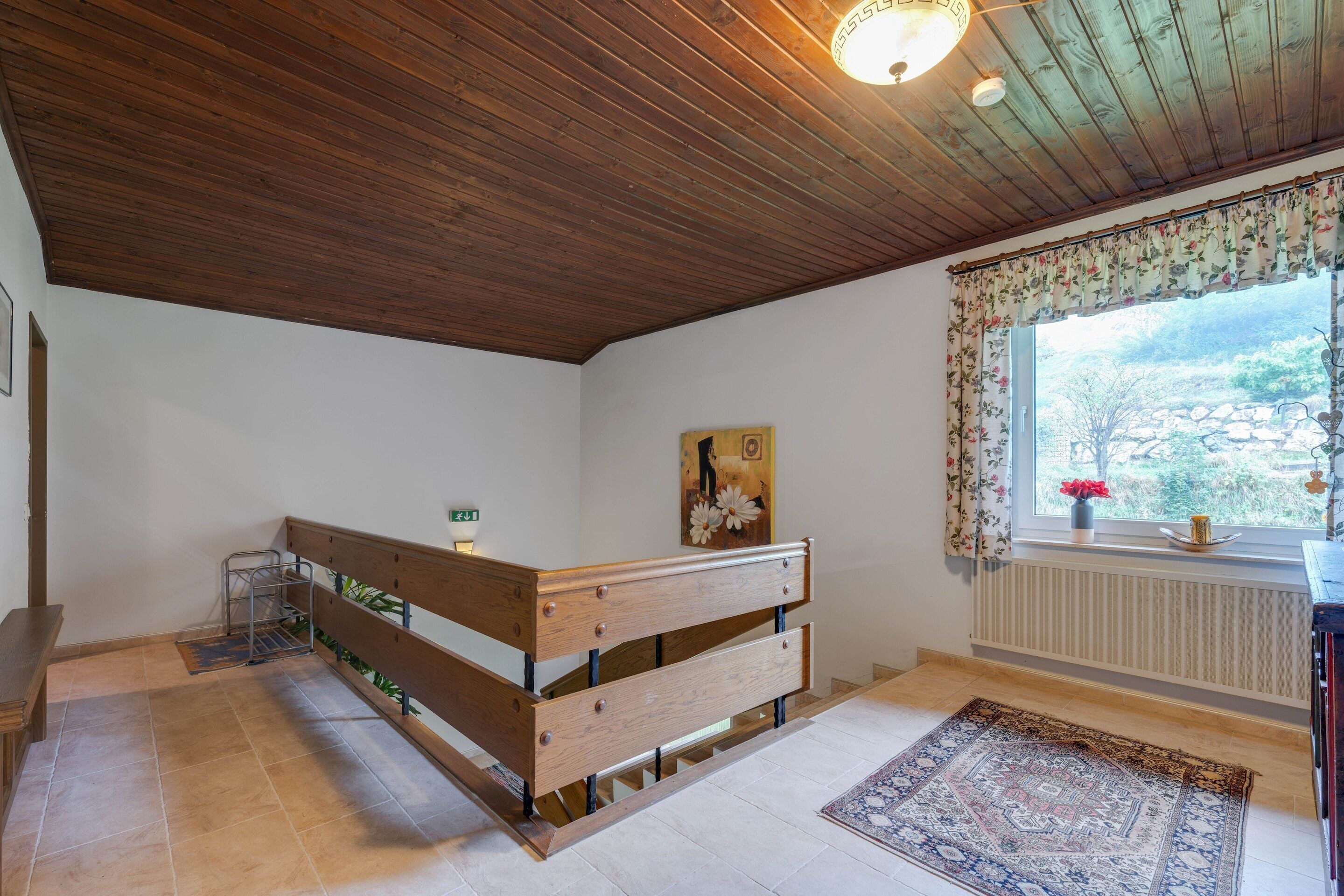 Een kamer met een houten bankje, een radiator, een raam met een bloemen gordijn, en een schilderij aan de muur.