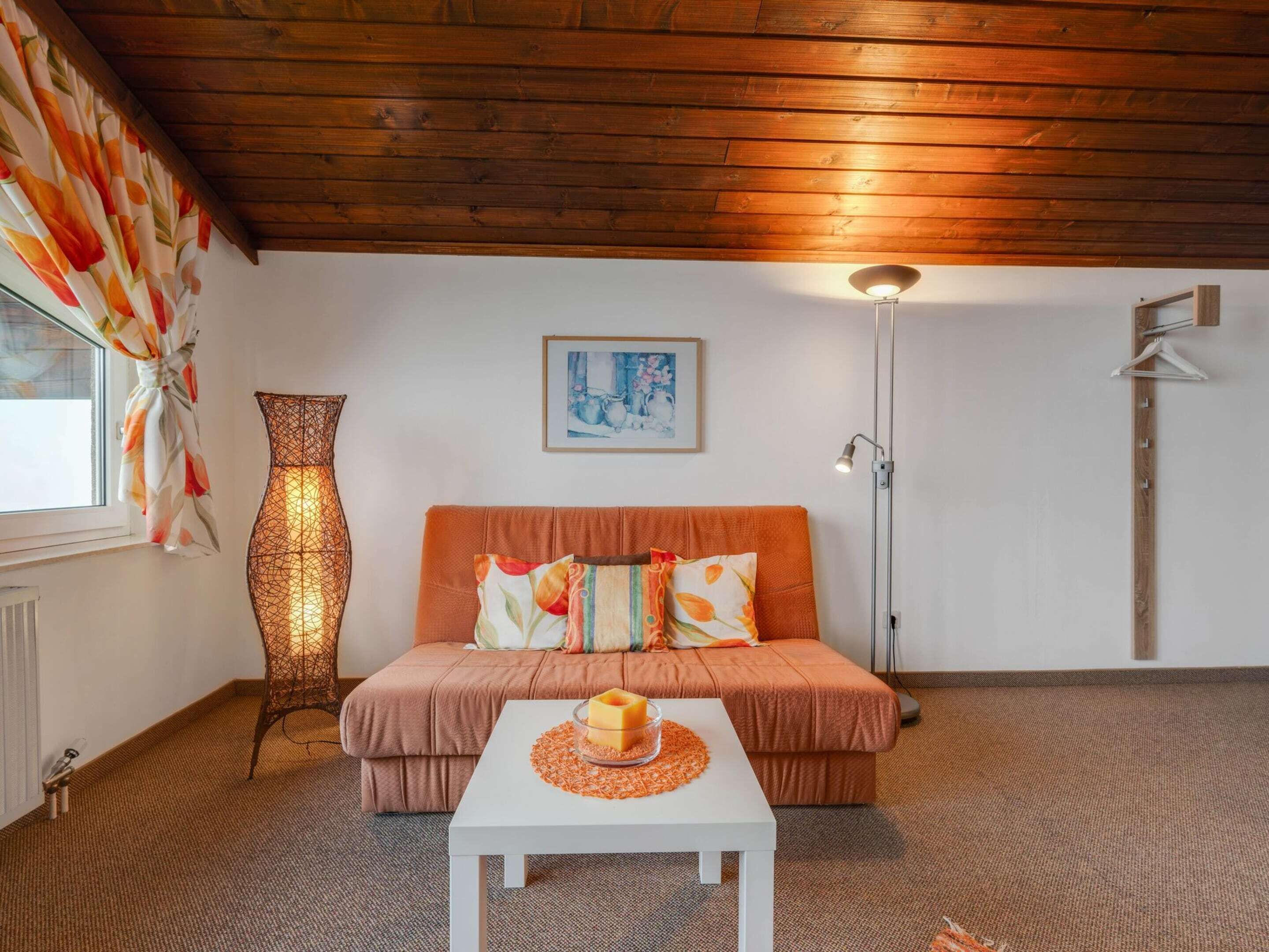 Un cuarto acogedor con techo de madera, un sofá con cojines anaranjados, una mesa blanca con un cuenco de vidrio, una lámpara de pie alta y un jarrón grande.