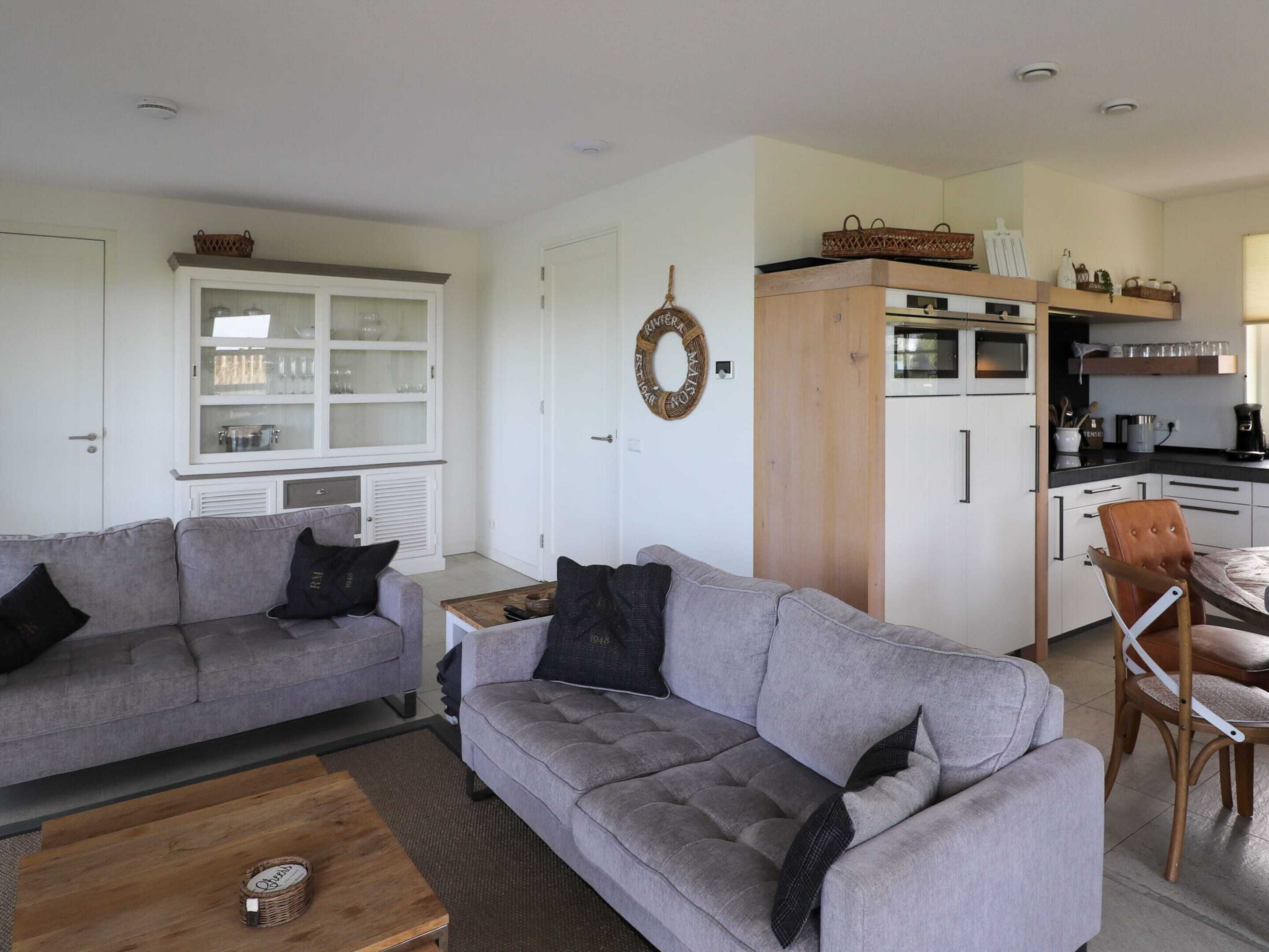 A modern living room with two grey sofas, a wooden coffee table, and a kitchen area with white cabinets and a wooden countertop.