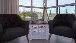 A modern living room with two dark armchairs, a white side table, and a floor lamp. Large windows offer a view of the outdoors. - Luxury Villa with Sauna & Hot Tub at Lake (Delfstrahuizen)
