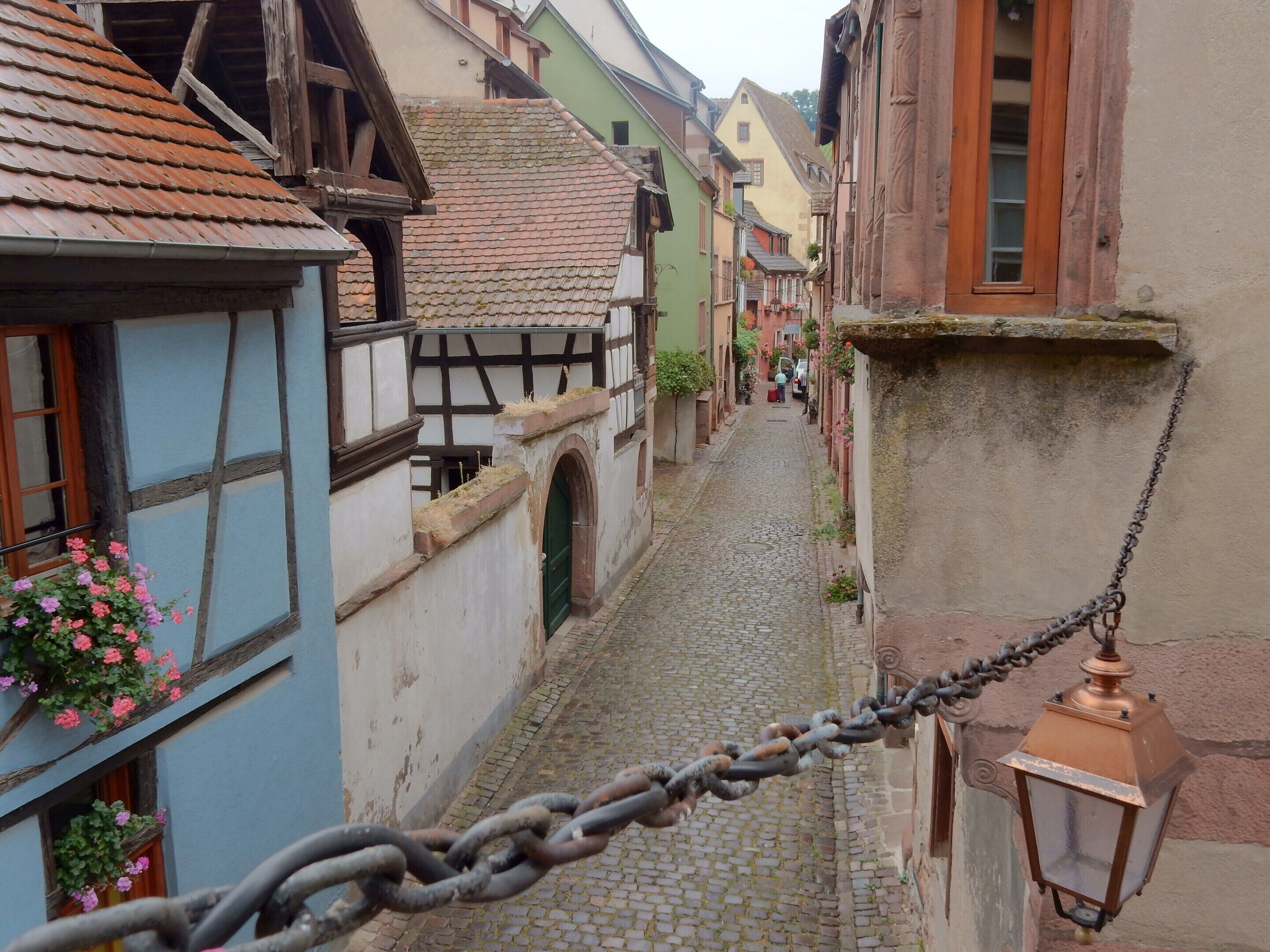 A cobblestone street flanked by traditional half-timbered buildings with a chain-link fence and a hanging lantern.