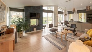 A spacious living area with a fireplace, a dining area, and a kitchen with large windows. - Chalet in Zeeland With Garden and Fireplace (Wemeldinge)