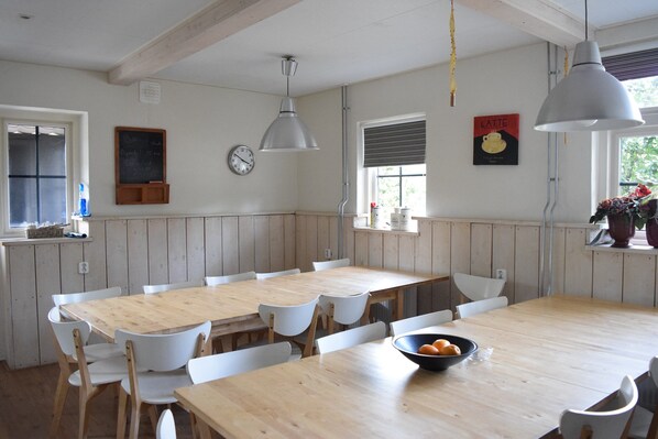 A dining area with wooden tables and white chairs, a wall clock, and a window with blinds. - Spacious Farmhouse with Barbecue near Veluwe (Voorthuizen)