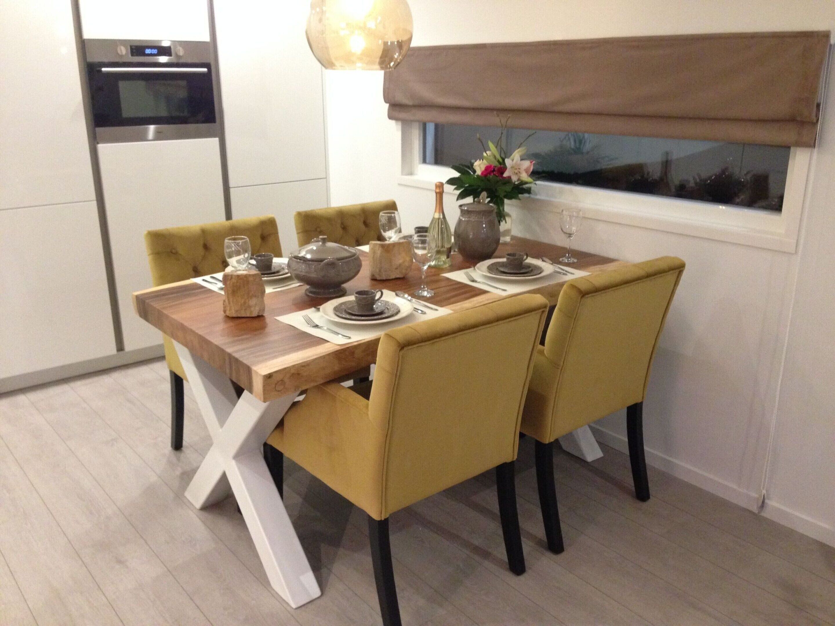 Een moderne eetruimte met een houten tafel voor zes personen, gele stoelen en een uitzicht op het buitengebeuren door een raam met bruin zonnescherm.