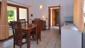 A dining area with a wooden table and chairs, a small refrigerator, and a kitchen visible through an open door. - Residence a Badesi 3377 (Badesi)
