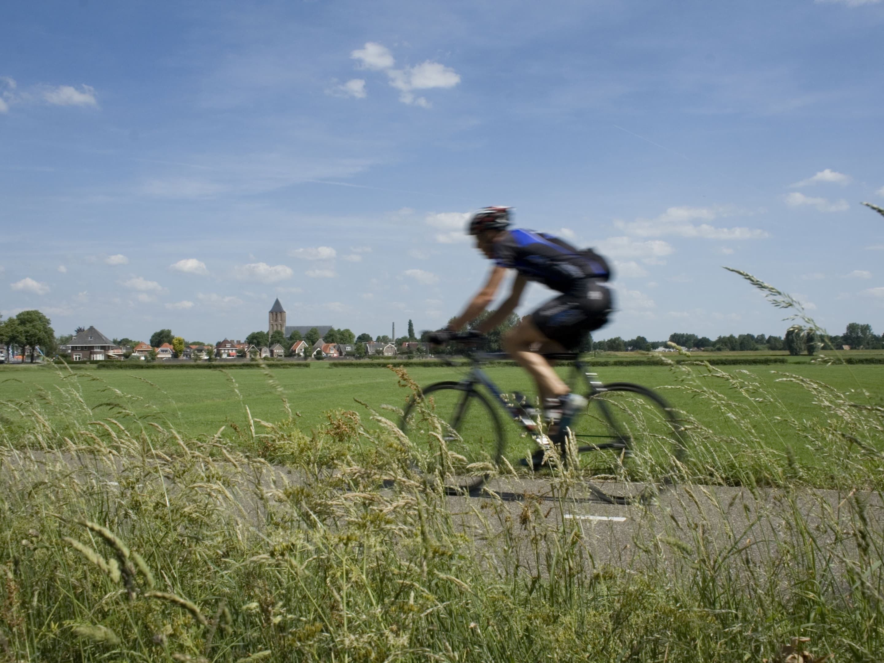 Un cycliste circule sur une piste asphaltée, avec un paysage rural en arrière-plan, comprenant des maisons et le clocher d’une église.