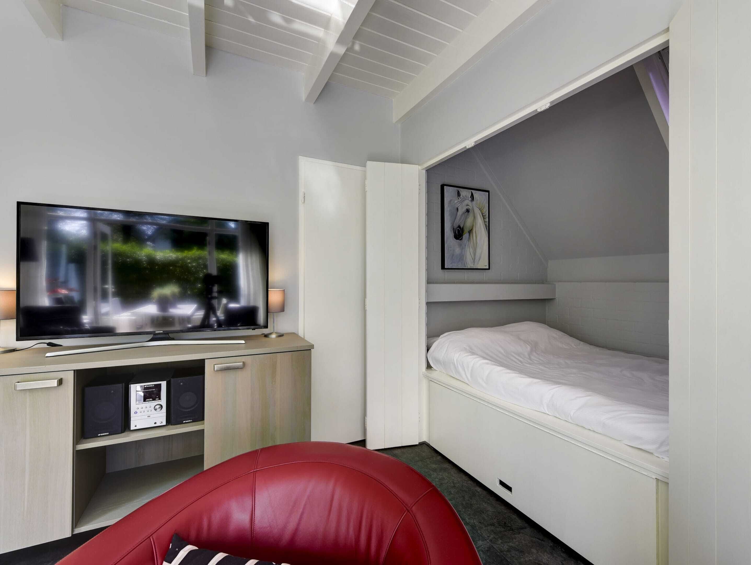 A compact room with a TV, a bed, a red chair, and a small desk with speakers.