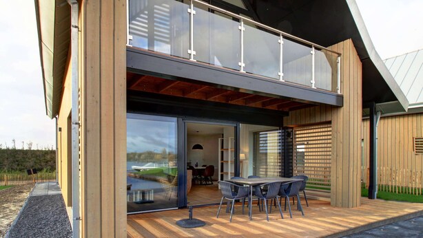 A modern two-story building with a large glass balcony, a dining area with outdoor seating, and a view of a body of water. - Villa Oesterdam Deluxe 6 Persons (Tholen)