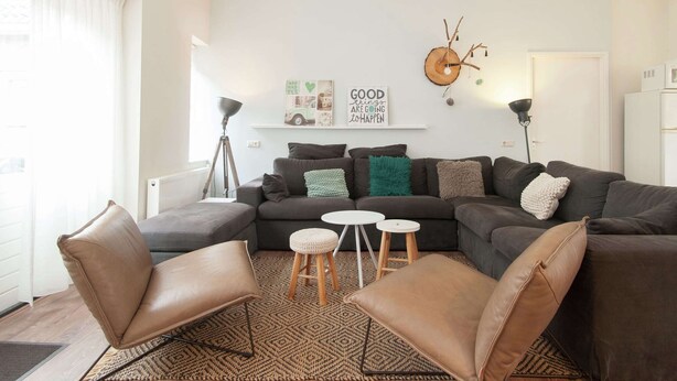 A modern living room with a grey sofa, two leather armchairs, and a small coffee table. There is a floor lamp, a wall-mounted shelf with decorative items, and a large window with sheer curtains. - Holiday Home in Bergen Near North Sea Beach (Bergen aan Zee)