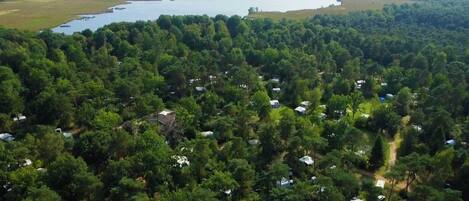 An aerial view of a campground with numerous RVs and tents, surrounded by dense forest and a large body of water in the background.