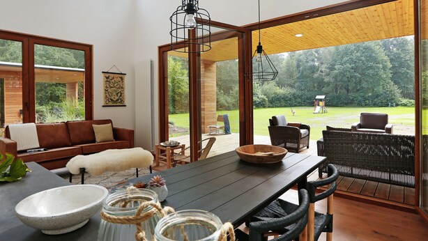 A spacious room with a large wooden table, wicker chairs, a sofa, and a view of a lush garden through large windows. - Bright & Airy Forest Cabin (Enter)