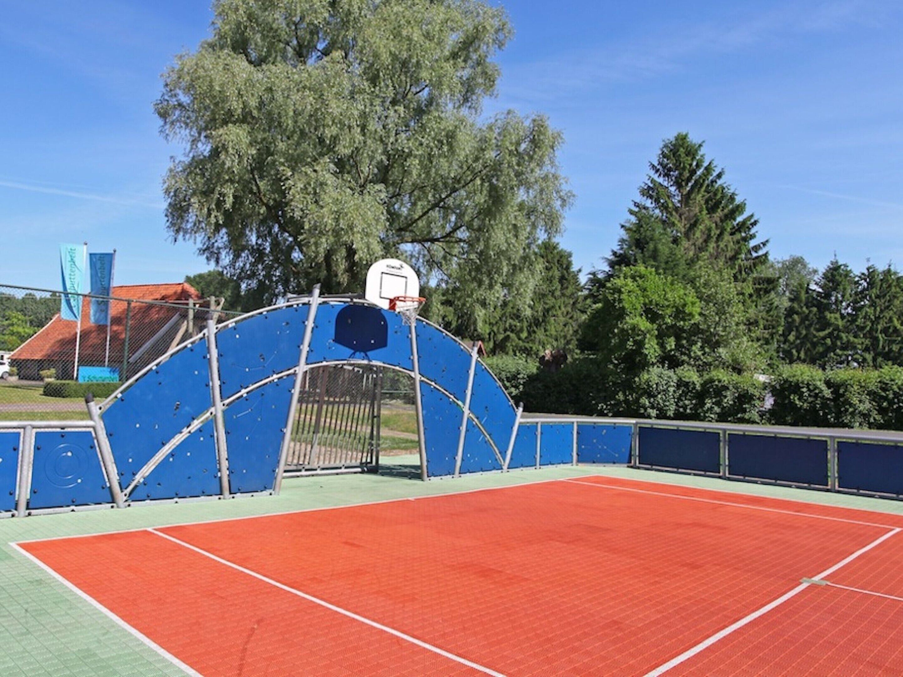 Una cancha de baloncesto al aire libre con superficie azul y roja, rodeada por una valla metálica y árboles.