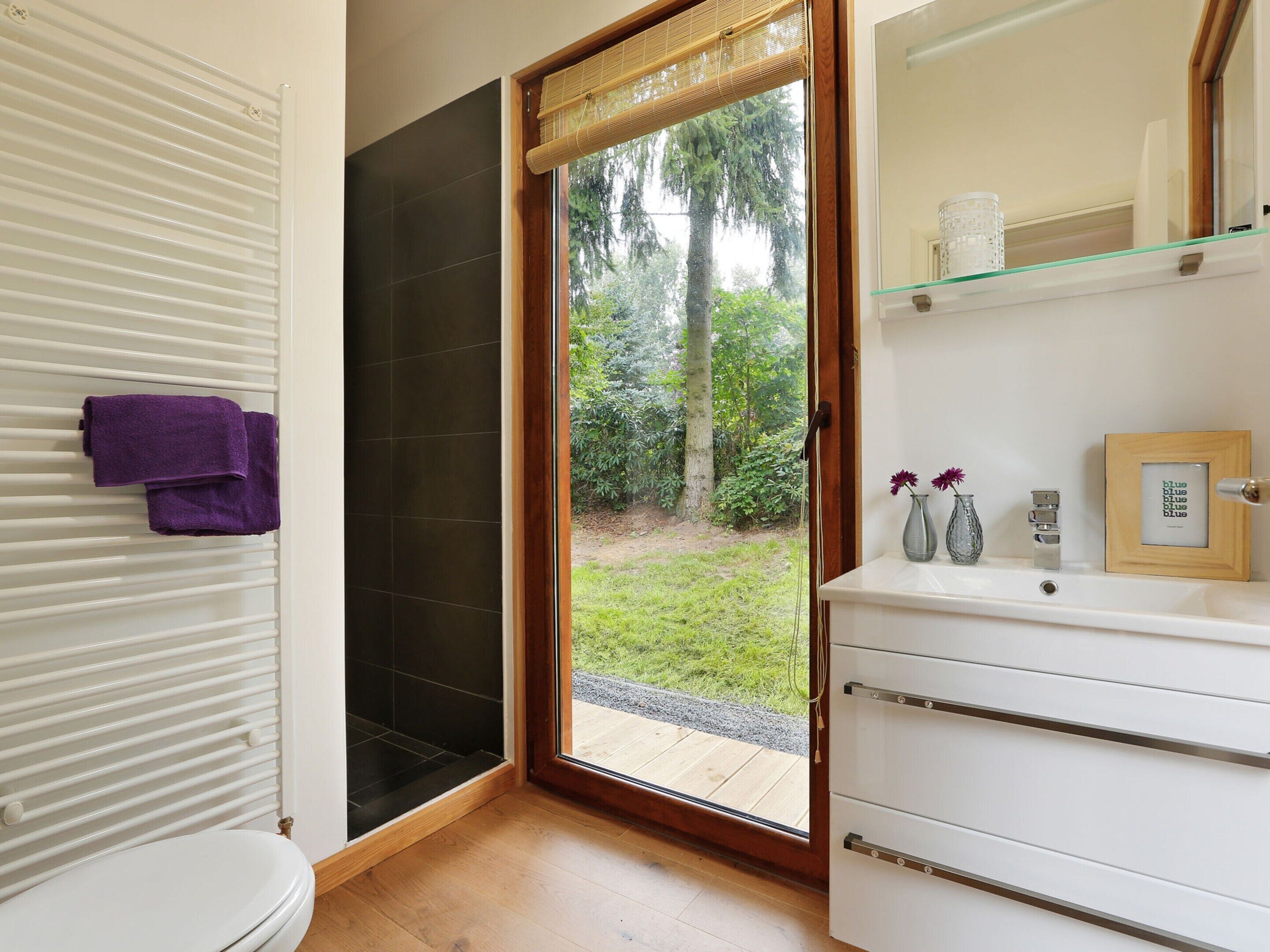 A bathroom with a toilet, a sink with a mirror, a shower area, and a view of greenery outside.