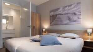 Une chambre à coucher avec un grand lit, une tête de lit en bois, une table de chevet avec une lampe, un tableau au mur et une salle de bain avec un miroir et des placards.