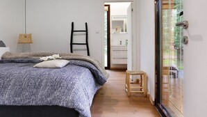 Een slaapkamer met een bed, een houten ladder, een klein krukje en een badkamer die door een open deur zichtbaar is.