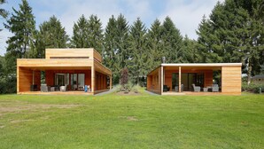 Een modern houten vakantiehuis met twee verdiepingen, grote glazen deuren en ramen, omgeven door een verzorgd gazon en dicht bos.
