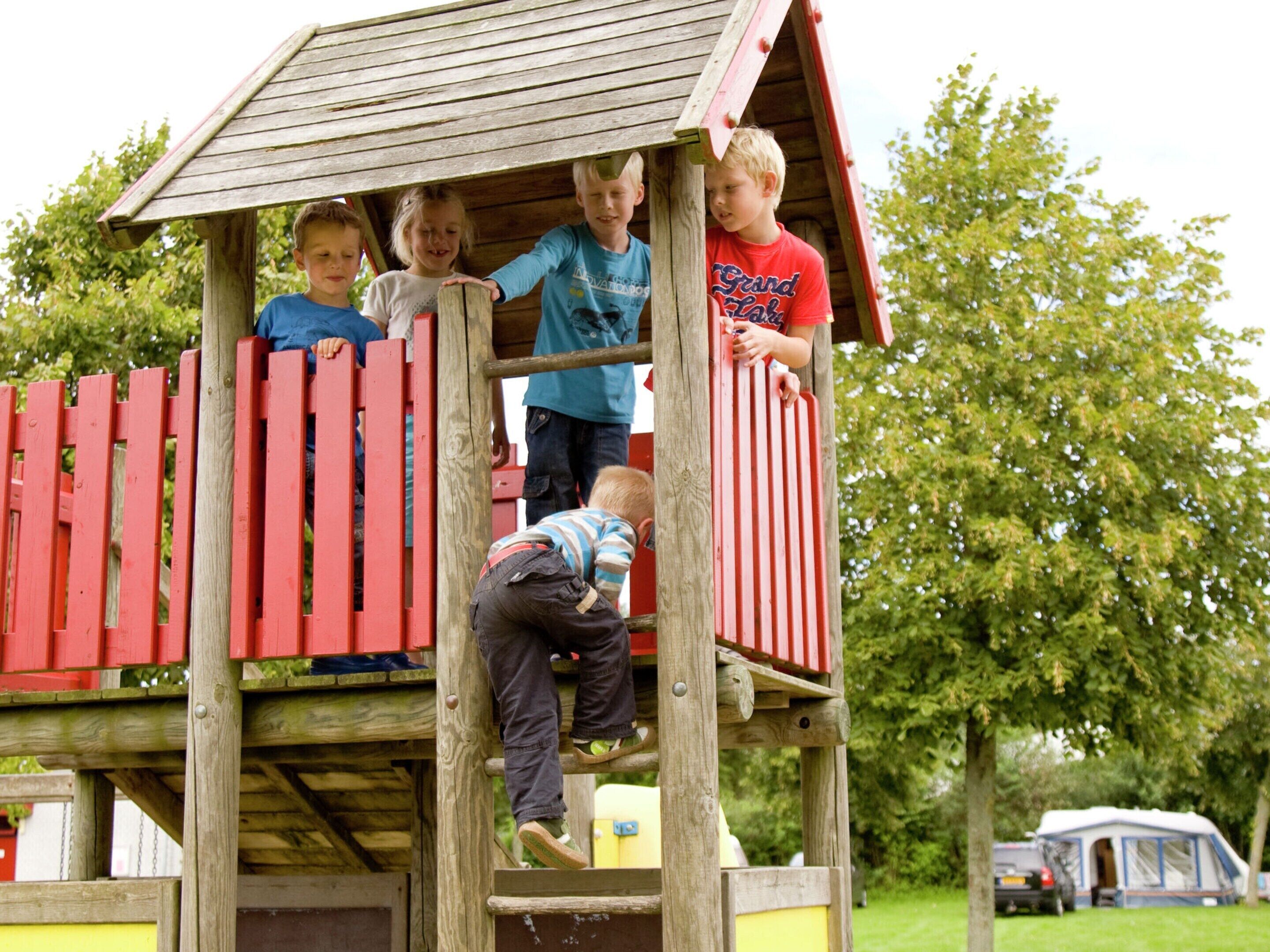 A wooden play structure with a red roof and a yellow base, surrounded by greenery.