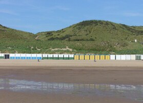 Una playa con una hilera de cabañas de playa coloridas, arena y una colina al fondo.