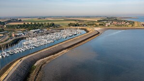 An aerial view of a marina with numerous boats docked along a narrow waterway, surrounded by fields and a small settlement.