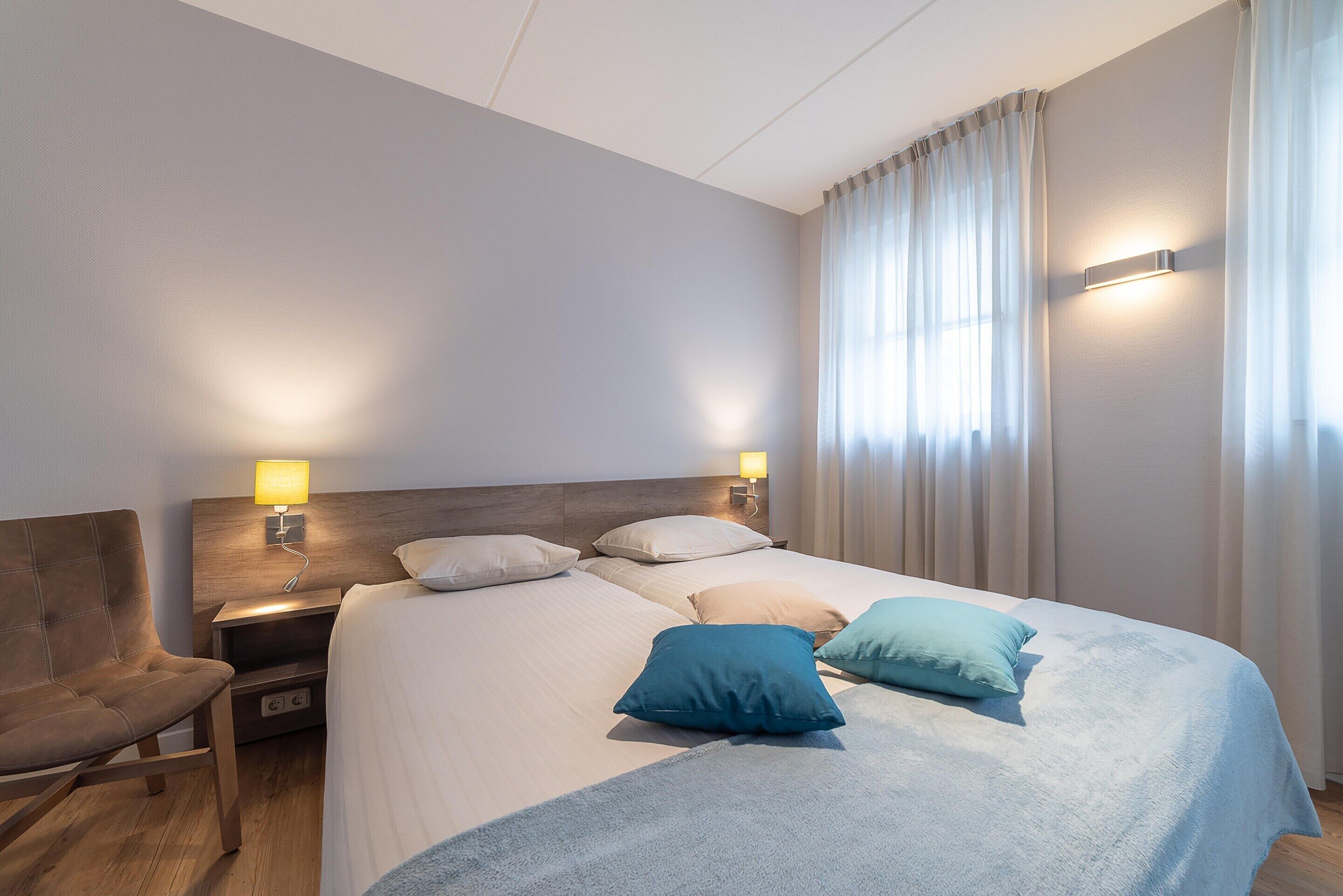 Ein Hotelzimmer mit einem Bett, zwei Nachttischen mit Lampen, einem Sessel und einem Fenster mit Vorhängen.