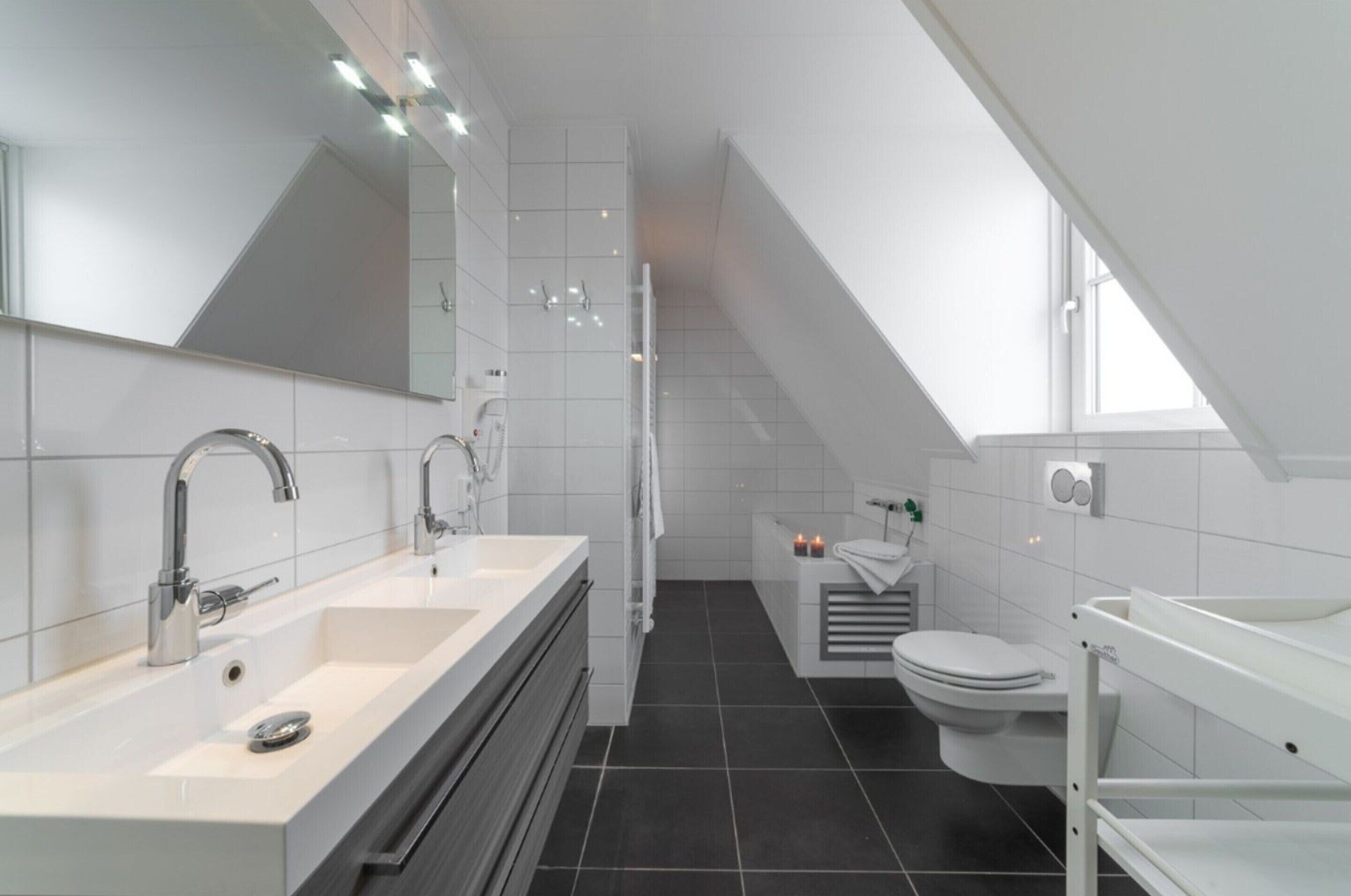 A modern bathroom with a large mirror, two sinks, a toilet, and a shower area.