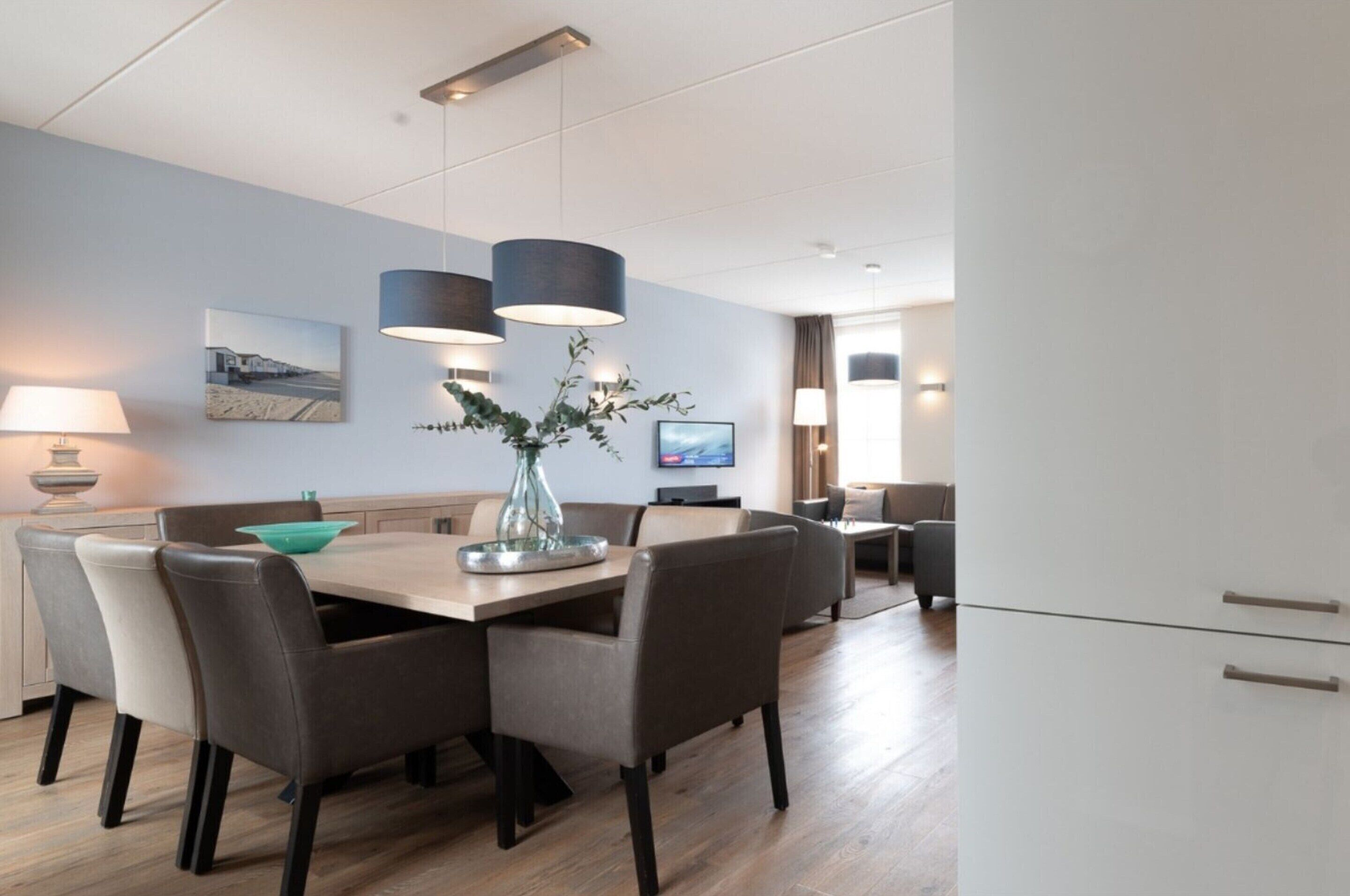 Un espace repas moderne avec une table en bois, des chaises et des luminaires suspendus. On y trouve également un téléviseur et un vase avec des plantes sur la table.