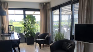 Un saló modern amb un sofà, una butaca i una televisió. Hi ha una finestra gran amb persianes, una planta en test i una zona de dinar amb taula i cadires.