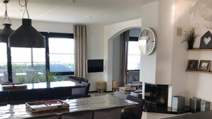 Un saló modern amb un sofà, taula de menjador i cadirees. Hi ha una televisió plana penjada a la paret, un rellotge i una finestra amb cortines.