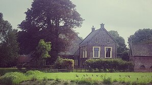 Uma casa tradicional com fachada de pedra, uma grande árvore na frente e um lago com patos.