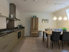 Moderni keittiö, jossa on keskeinen saareke, sisäänrakennettu uuni ja ruostumattomasta teräksestä valmistetut kodinkoneet. Keittiössä on ruokailutila, jossa on puinen pöytä ja tuolit, ja pöydällä on pieni ruukkukasvi.