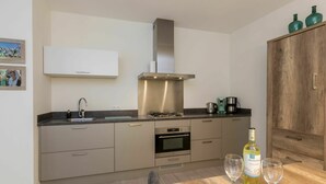 Een moderne keuken met een roestvrijstalen afzuigkap, een inbouwoven en een koffiezetapparaat. Aan de rechterkant staat een houten kastje en een eettafel met twee wijnglazen en een fles.