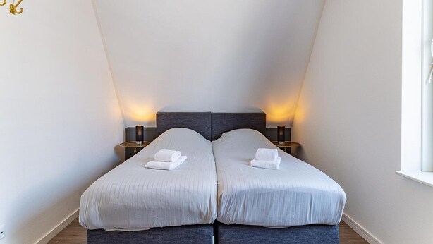A bedroom with two beds, each with a white towel, two bedside tables with lamps, and a window on the right. - Apartment Near South Beach With Sauna (Koudekerke)