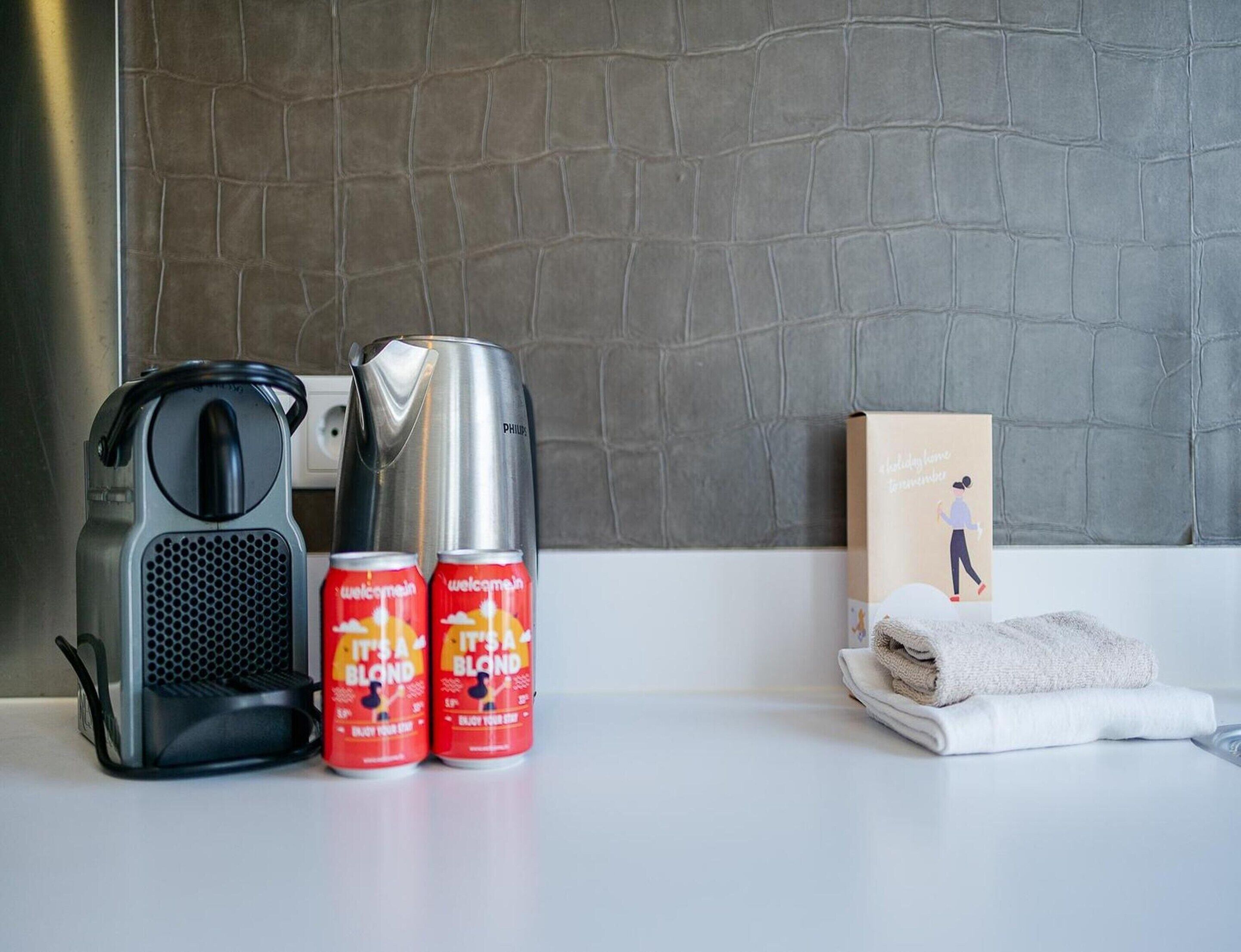 A kitchen counter with a coffee maker, a kettle, two cans of 'It's a Blond' beverage, a towel, and a box with a person illustration.