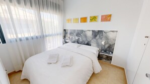 Ein Schlafzimmer mit einem Bett, weißen Vorhängen, einer Wand mit abstrakter Kunst und einem gefliesten Boden.