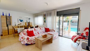 Ein modernes Wohnzimmer mit einer Couch, einem Holztisch, einem Essbereich mit Stühlen, einer Küchenzeile und einer Glastür zum Pool.